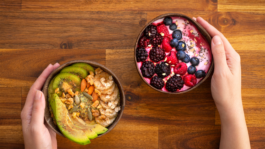 Two bowls filled with healthy snacks on the go, on a wooden table, being grabbed by a set of hands.