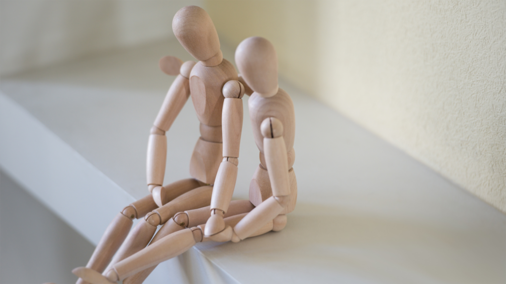 Two artist mannequins posed to show that one is comforting the other with its arm around the second mannequin's shoulder, both sitting on a white shelf, representing Healthy Communication in Relationships.