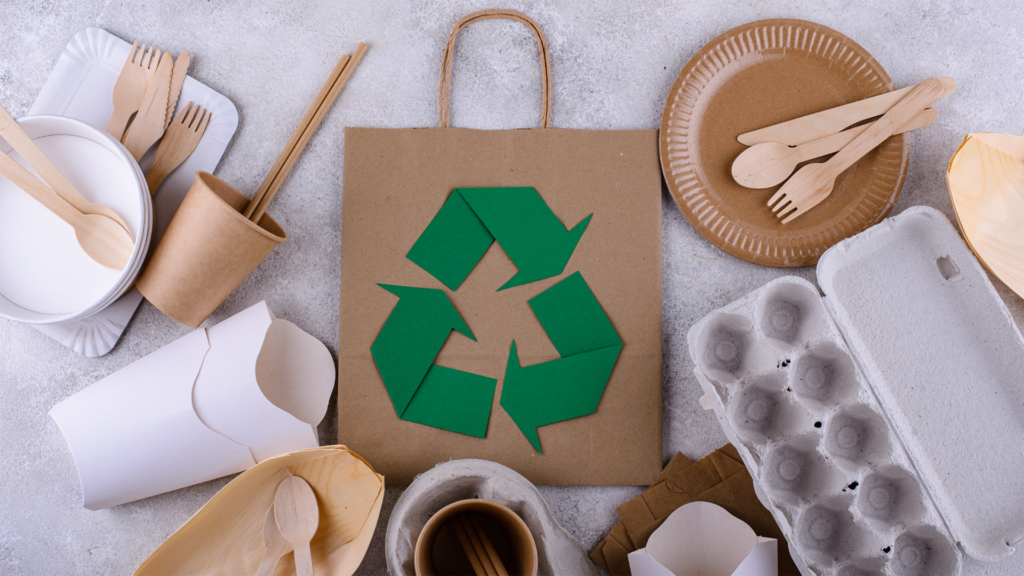 A collage of various different recyclable materials around a centerpiece of a kraft bag with cut-out paper shapes that make up a recycle icon, illustrating how it's become easier to live a green lifestyle by recycling.