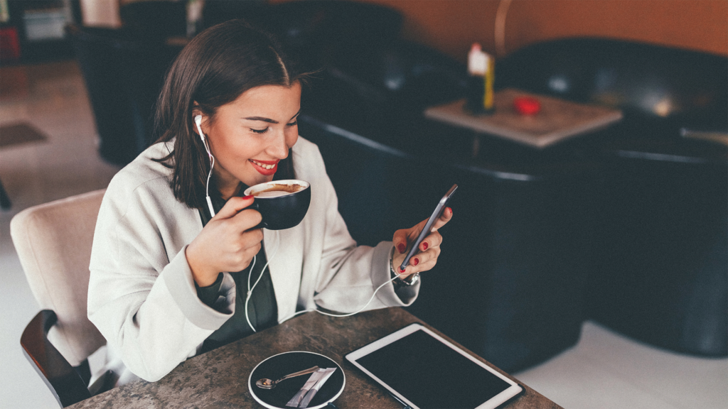 A young woman drinking a latte in a coffee shop at a table with a tablet device and headphones plugged in to a phone, smiling at the phone screen as if she is reading something amusing, showing how to stay in touch with friends even while living far away.