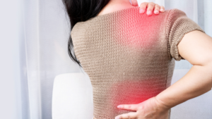 A woman with dark hair in a tan sweater holding reaching back to massage painful areas in her upper and lower back, as illustrated by areas highlighted in red - how to relieve body aches.